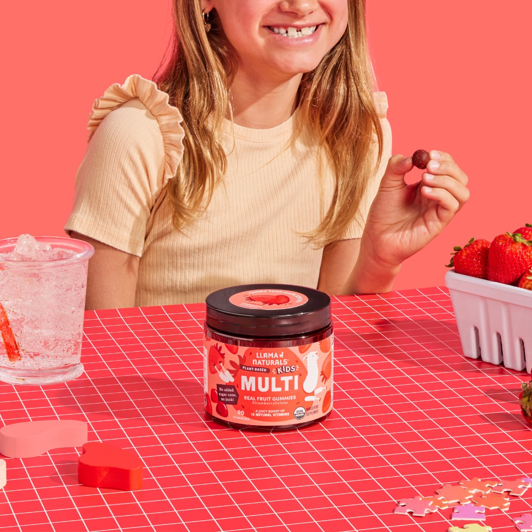 Child at a kitchen table reaching for a Llama Naturals Kids Multi gummy, surrounded by strawberries