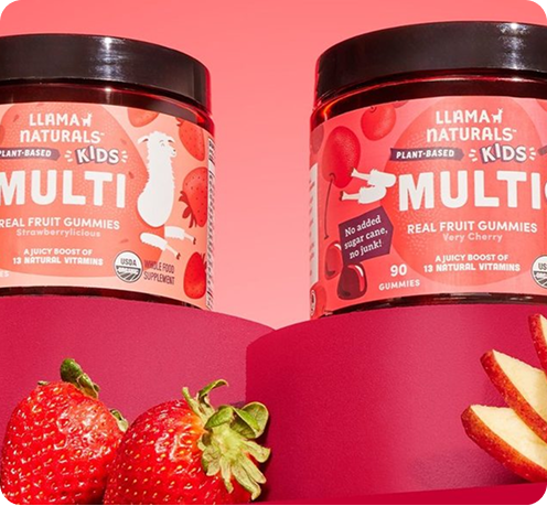 Two Llama Naturals Kids Multi jars surrounded by strawberries, apple slices, oats, and kale